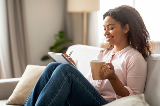 Woman journaling at a desk while holding a mug, surrounded by cozy workspace items, promoting mindfulness and mental wellness.